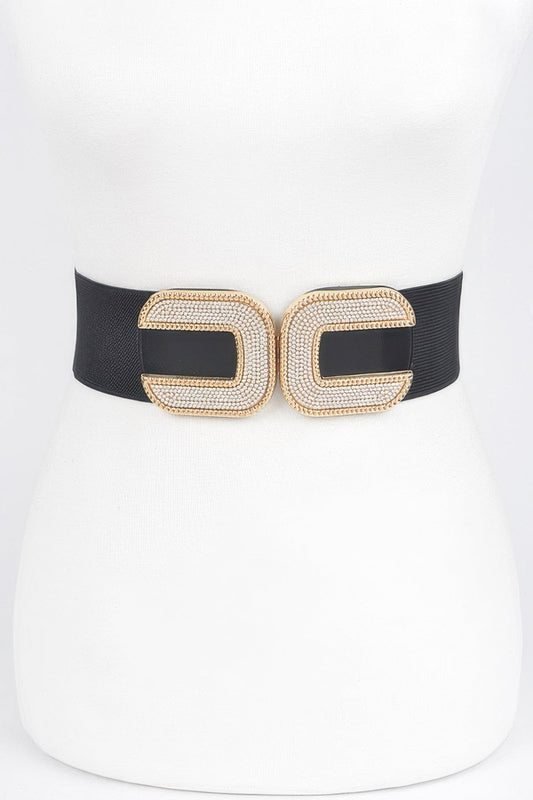 Front view of a black elastic stretch belt featuring a double interlocking gold-tone clasp encrusted with sparkling rhinestones, styled around a white dress mannequin against a light background.