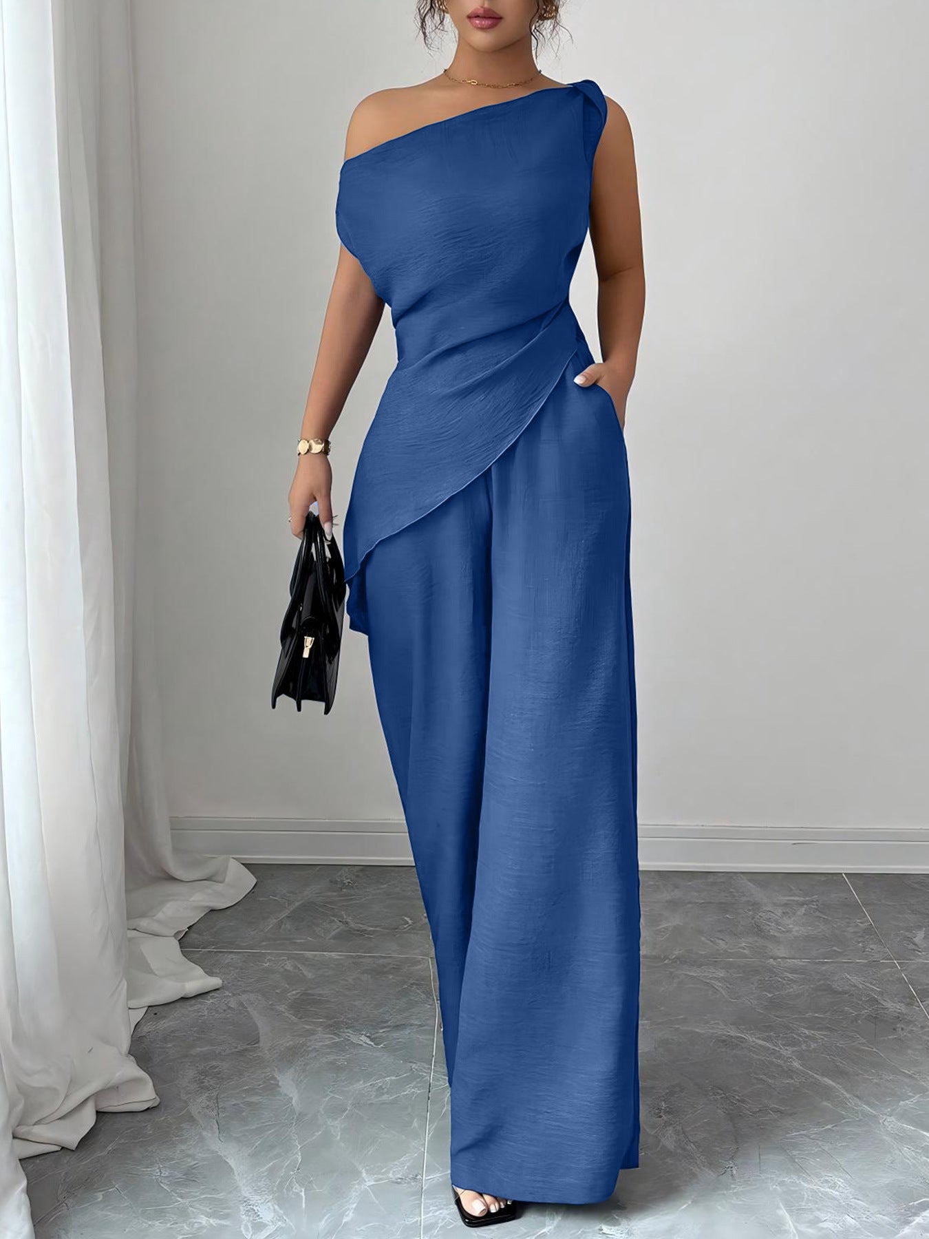 Woman wearing a royal blue casual two piece set featuring an asymmetric one-shoulder top with a diagonal hem and matching high-waisted wide-leg pants, holding a black handbag.