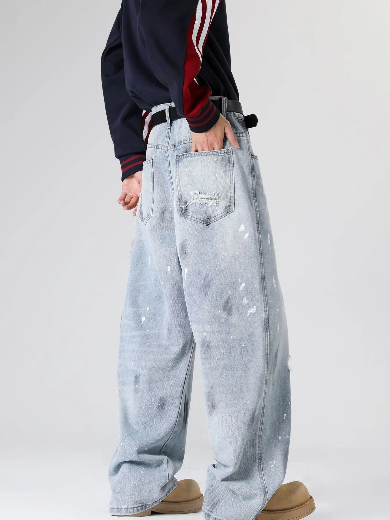 Rear-view of a person wearing light blue distressed wide-leg jeans with paint splatter details, highlighting the back pockets, belt, and dark sweatshirt against a dark background.
