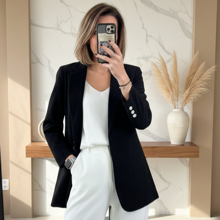 Woman wearing a black slim-fit corduroy blazer over a white top and white trousers, standing indoors next to a decorative vase.