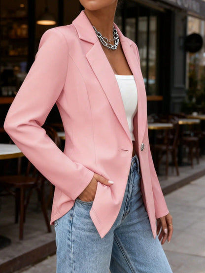 Woman wearing a pink blazer, white top, and blue jeans, standing outdoors on a city sidewalk beside a café.