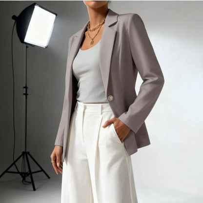 Woman wearing a taupe blazer, white top, and white wide-leg trousers, posing in a professional studio setting with a softbox light visible in the background.
