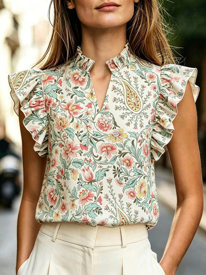 Woman wearing a floral and paisley print sleeveless top with a high ruffle-trim mock neck and lotus-leaf ruffle sleeves, styled with white high-waist pants.