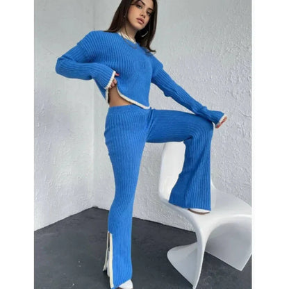 Woman wearing a vibrant blue ribbed knit long-sleeve crop top and matching split-hem pants with white trim, standing with one foot on a white modern chair.